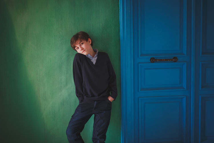 A serious cute teen boy with blue eyes is standing by the green wall. Next to a bright blue door. Pensive Blue Boy Casual Clothing Childhood Contemplation Door Front View Green Color Indoors Lifestyles Looking Looking Away One Person Real People Standing Teenager Three Quarter Length Young Adult