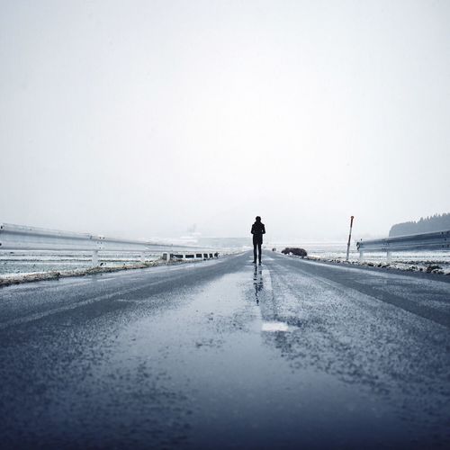 Full length of person standing on wet street amidst snow covered field against clear sky