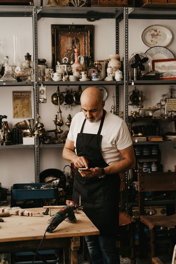 Serious middle aged male carpenter in apron messaging on mobile phone while standing near workbench with professional tools in creative work studio