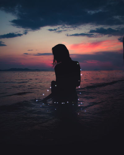Silhouette woman with illuminated string lights in sea against sky during sunset