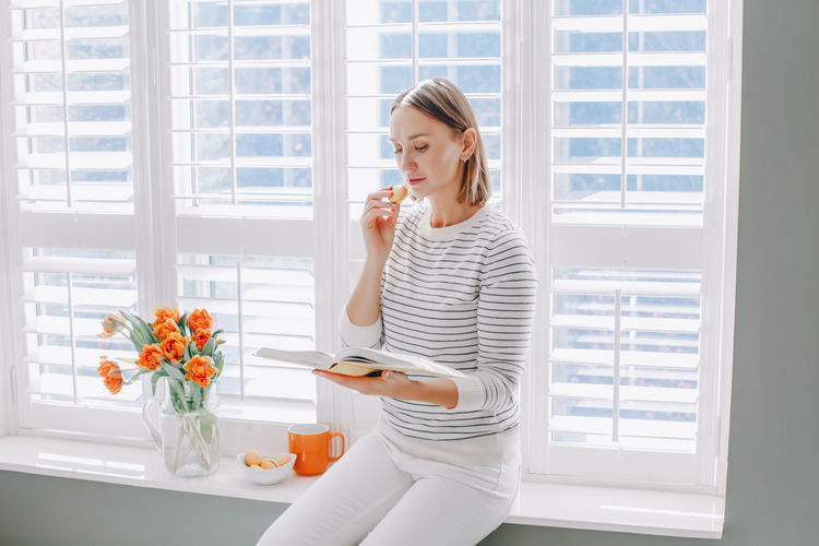 Woman eating food while reading book sitting by window at home