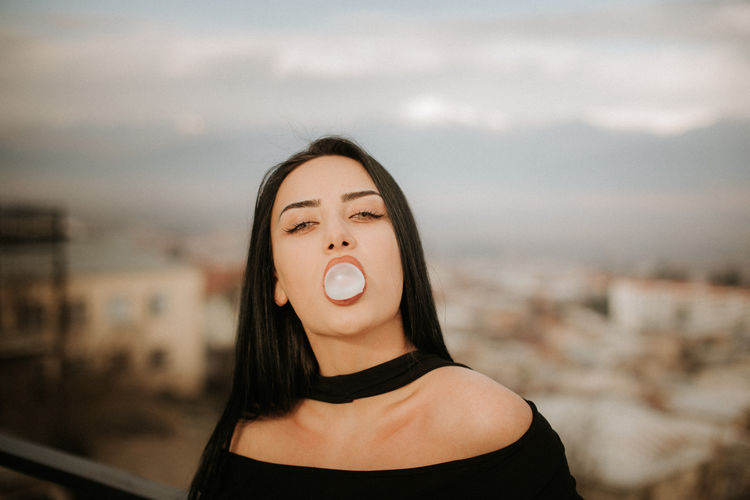 Portrait of beautiful young woman chewing gum against cityscape