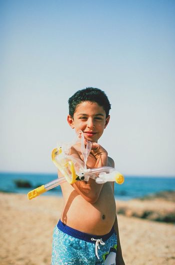Portrait of smiling cute boy holding scuba mask while standing on sand at beach