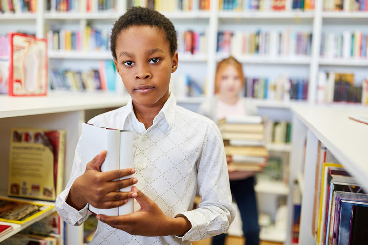 Portrait of boy holding book while standing in school library