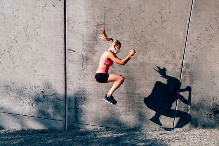 Fit female athlete doing squat jump outodoor against a gray wall. sporty woman wearing face mask