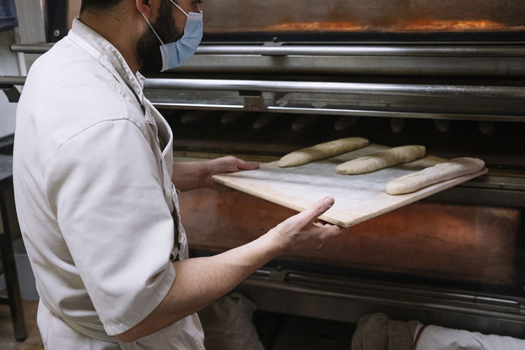 Male chef putting bread dough in oven at bakery during covid-19