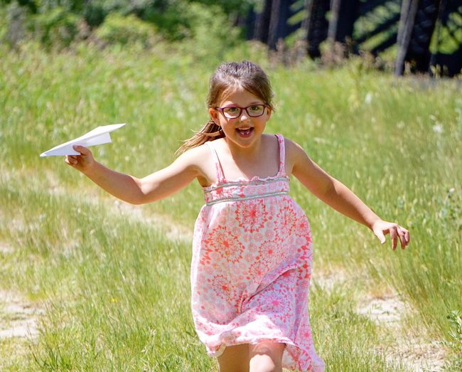 Cheerful girl flying airplane while walking on field during sunny day
