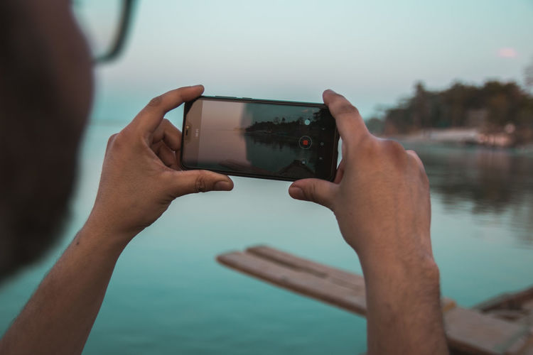 Cropped hands of man photographing lake with smart phone