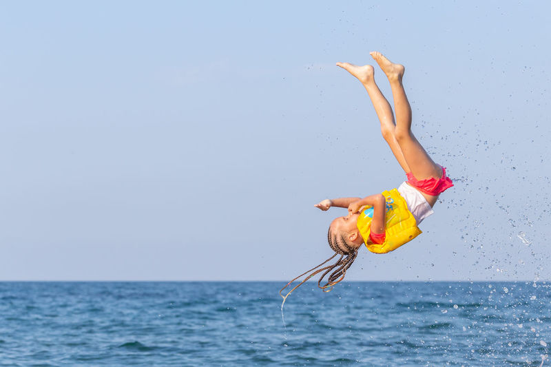 Little girl with 4-5 years in yellow life jacket flies upwards against background of blue sky, sea.