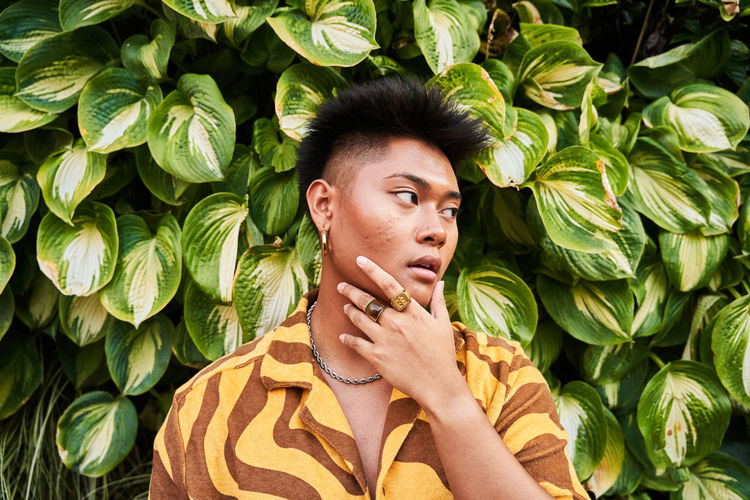 Dreamy thoughtful young filipino non binary man wearing cool shirt with earring standing in green garden looking away