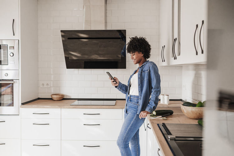 Happy woman using smart phone leaning on kitchen counter at home