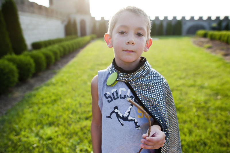 Portrait of boy wearing cape and holding stick while standing on grassy field at park