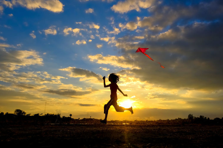 Silhouette girl running while flying kite in sky during sunset