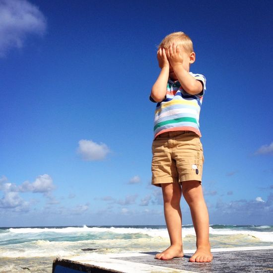 Low angle view of boy covering eyes while standing at sea shore against sky
