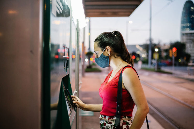 Side view of young female passenger in protective mask buying ticket for public transport on vending machine