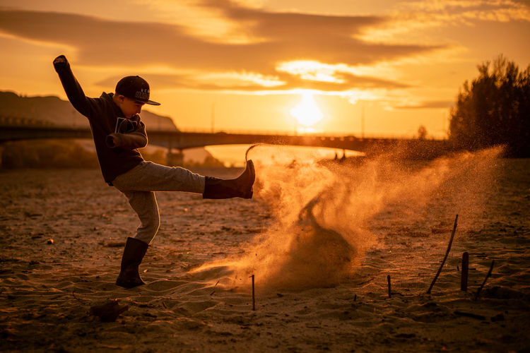 Full length of boy kicking sand on beach against sky during sunset