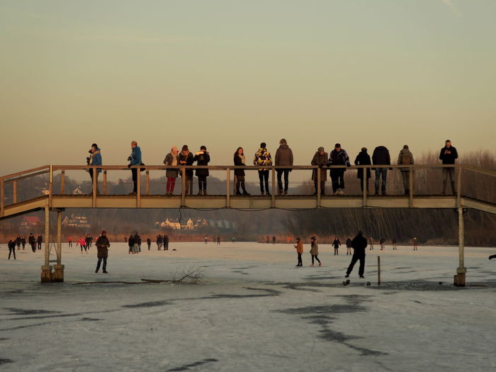 People on bridge over frozen lake against sky during sunset