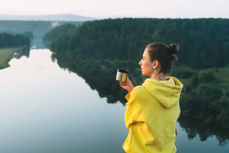 Rear view of woman holding coffee cup standing by lake