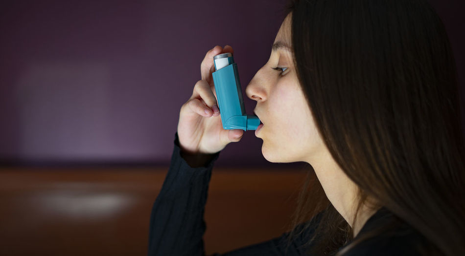 Woman inhaling a blue asthma inhaler at home.