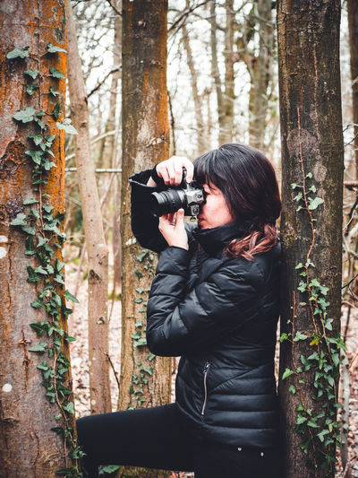 Full length of woman photographing through tree trunk in forest