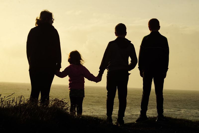 Silhouette woman with children standing on cliff by sea at sunset