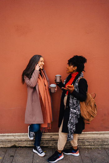 Side view of african american woman and friend in outerwear standing against red wall with takeaway hot drink and smartphones