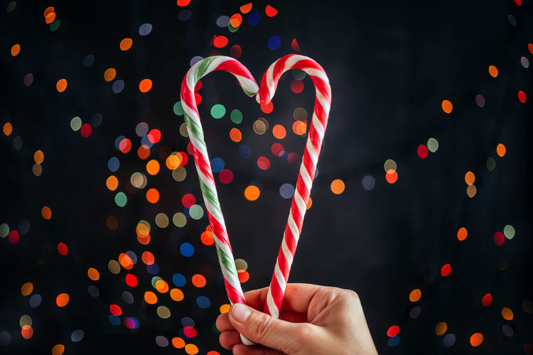 A hand holding candy canes in the shape of heart against christmas tree lights bokeh