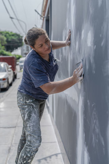 Side view of professional female painter in process of plastering wall using special equipment on city street