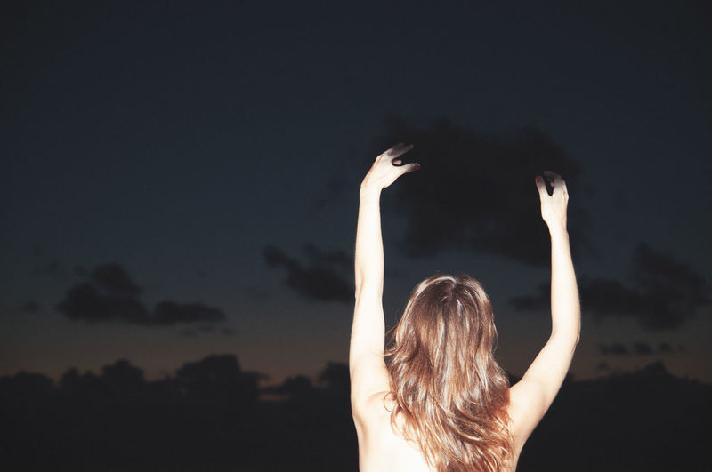 Rear view of woman with arms raised standing against sky at night
