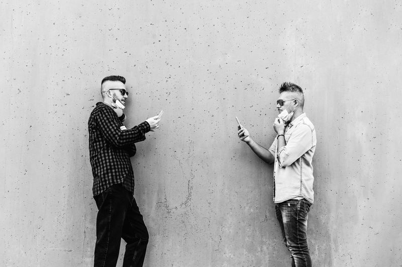 Full length of men holding mobile phone standing against wall outdoors