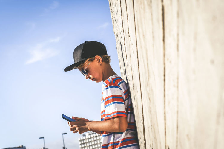 Side view of boy using phone while standing by wall