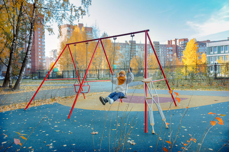 Child swinging on swing in playground with crutches in autumn. girl has one leg broken in cast.