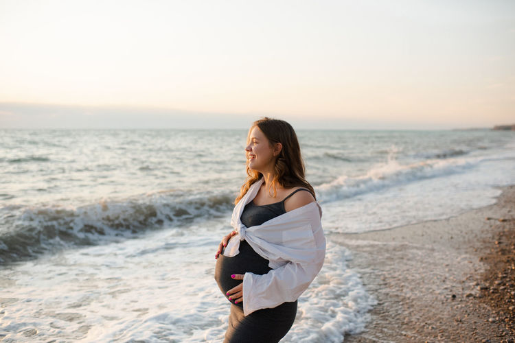 Smiling happy pregnant woman wear casual dress and white shirt standing over sea water outdoor.