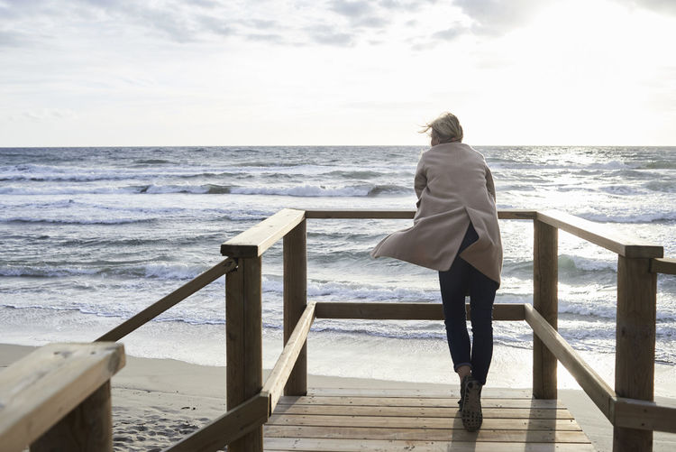 Spain, menorca, back view of senior woman standing on boardwalk in winter looking at the sea