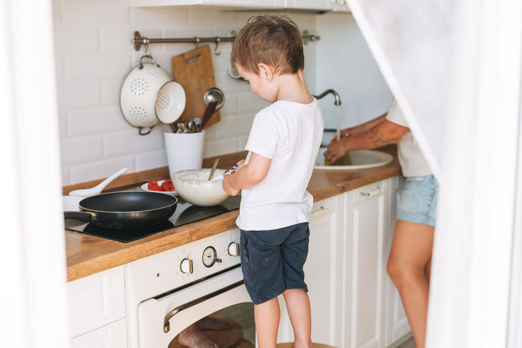 Cute toddler boy with mother cooking breakfast with puncakes together in bright kitchen at home