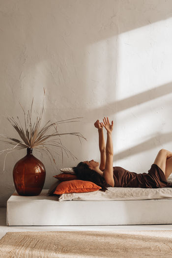 Young brunette woman in a silk brown dress lying on the bed in a cozy home interior with dry plants 