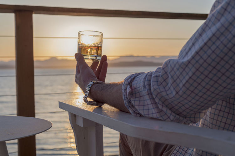 Midsection of man holding alcoholic drink while sitting by sea during sunset