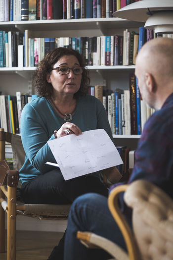 Mature female therapist showing analysis to patient against bookshelf at home office