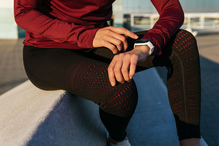 Crop anonymous female athlete in stylish sportswear sitting on border and checking pulse on fitness tracker while resting during outdoor workout in city