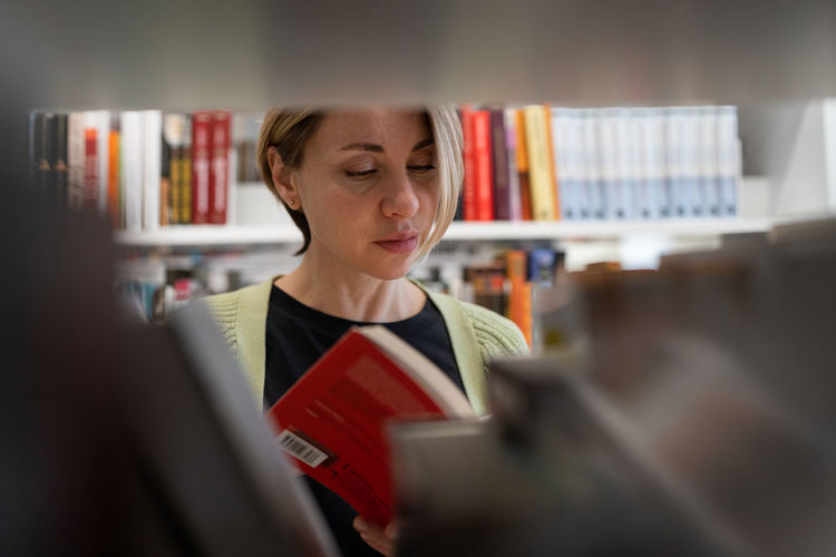 Female mature student looking for book at library bookshelf studying at university campus or college