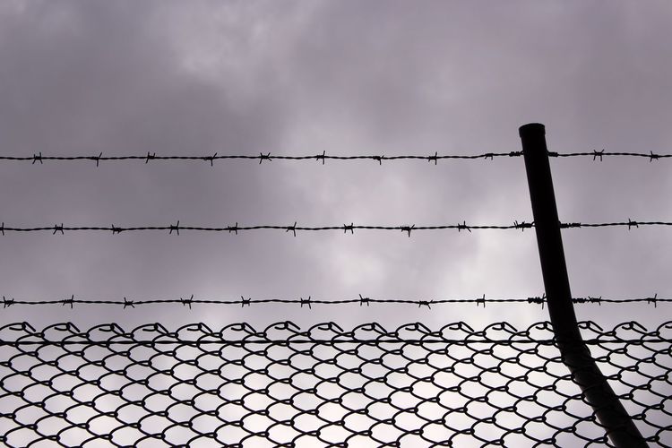 Silhouette of chain-link fence with barbed wire against cloudy sky