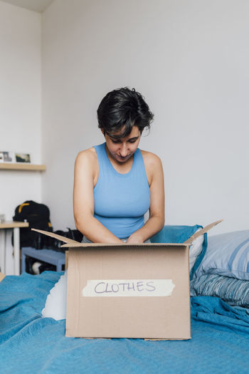 Young woman packing box for clothes donation on bed at home