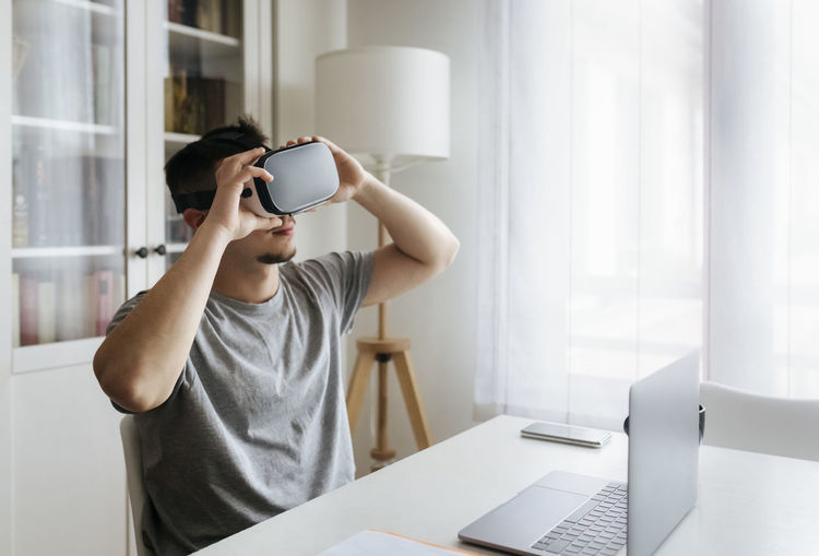 Male entrepreneur holding virtual reality headset while sitting with laptop on desk at home office
