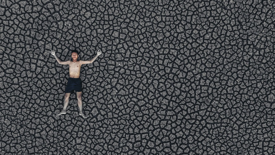 High angle view of shirtless boy with arms outstretched lying on cracked land