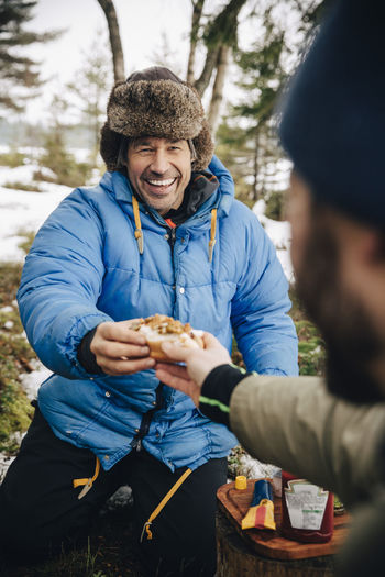 Cheerful man in warm clothing receiving hot dog from male friend