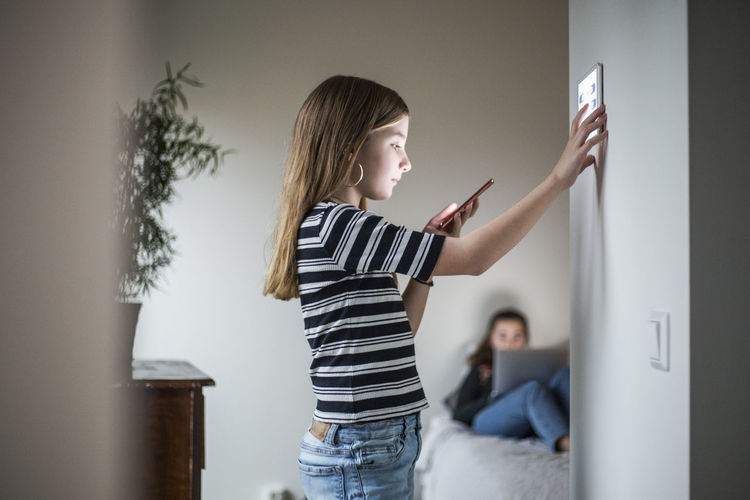 Girl looking at smart phone while using digital tablet on wall with sister in background at smart home