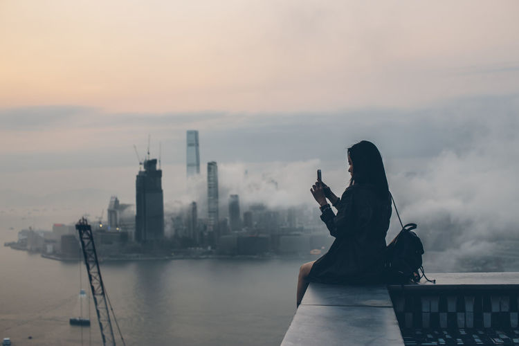 Woman sitting on retaining wall against sky in city