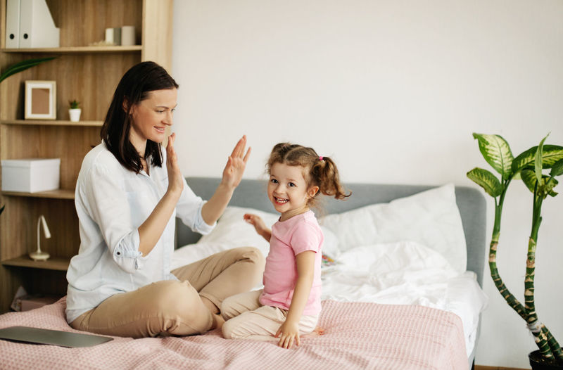 A young mother and a lovely daughter smile and clap their hands on the couch at home