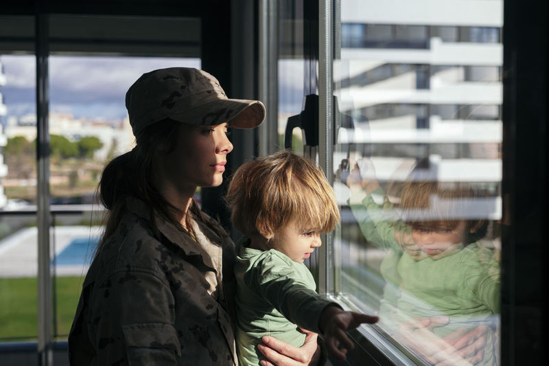 Side view of female soldier in uniform looking out window with cute son while standing in light room with sunlight