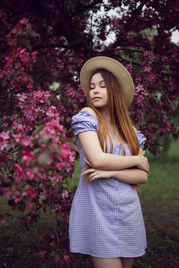 Portrait young beautiful girl teenager in a purple dress and in hat stands blooming pink apple tree